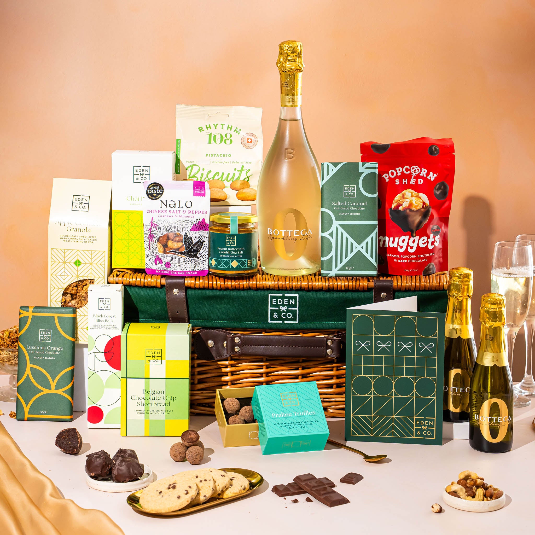 EDEN & CO. Luxury Wicker Hamper filled with a full-size Bottega Zero Black prosecco (0% alcohol-free), an assortment of chocolate bars, Booja-Booja Chocolate Wonders truffles and Buttermilk snack bars, presented in a dark green trimmed wicker basket with an “Occasion Themed” card, styled against a warm terracotta background.