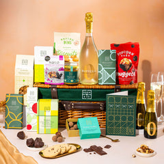 EDEN & CO. Luxury Wicker Hamper filled with a full-size Bottega Zero Black prosecco (0% alcohol-free), an assortment of chocolate bars, Booja-Booja Chocolate Wonders truffles and Buttermilk snack bars, presented in a dark green trimmed wicker basket with an “Occasion Themed” card, styled against a warm terracotta background.
