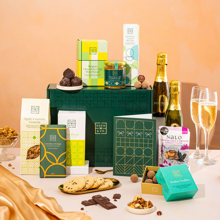 EDEN & CO. Food & Drink Hamper with a full-size Bottega Zero Black prosecco (0% alcohol-free), dark green gift cylinder, an “Occasion Themed” card, an assortment of chocolate bars, Booja-Booja Chocolate Wonders truffles, Buttermilk snack bars, a scented candle and Aery hand cream, styled with gold silk against a warm terracotta background.