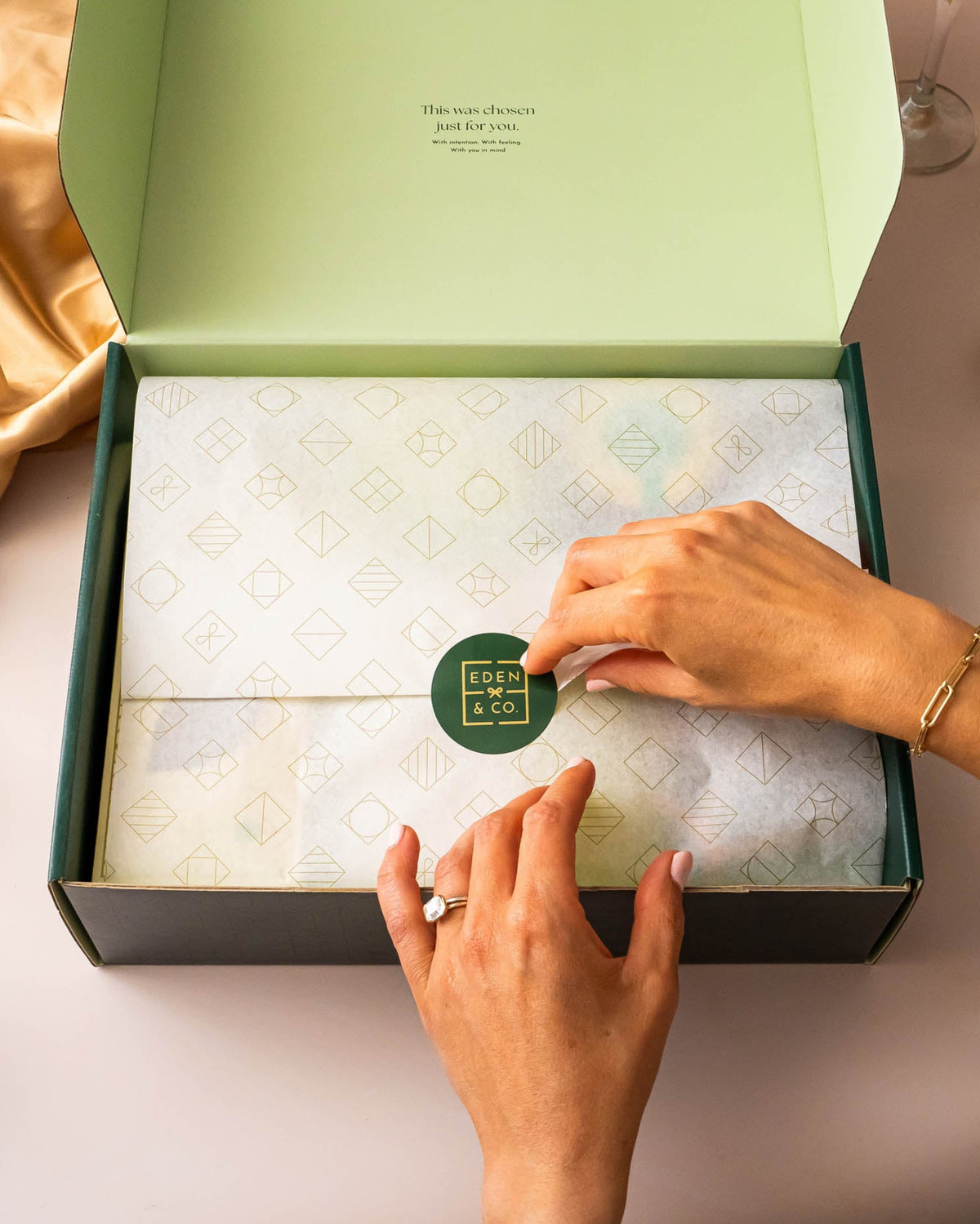 An open dark green gift box with a pale green interior, wrapped in patterned tissue paper and sealed with a round green sticker. Two hands are visible pressing the sticker, with champagne silk and a champagne bottle in the background.