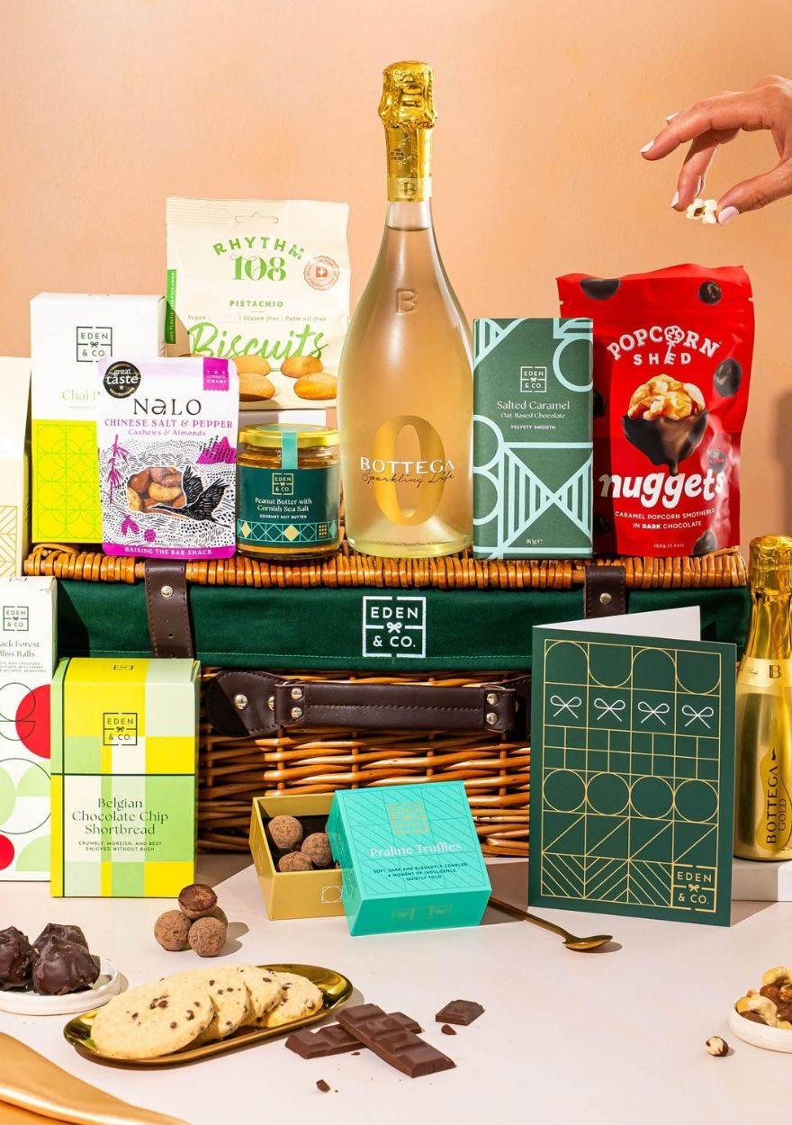 A gourmet gift hamper arrangement featuring a green Eden & Co basket filled with assorted packaged snacks, chocolates, and a bottle of Bottega Veneta sparkling wine, styled on a table with plates of cookies and chocolate pieces against a warm peach background. A hand is visible reaching in from the top right.