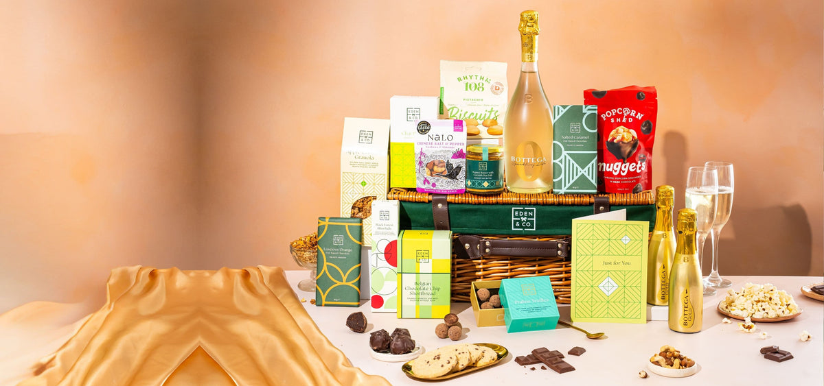 A luxury gourmet gift hamper arranged on a table against a warm beige background. The hamper includes a bottle of champagne, a bag of Nuggets, boxes of artisanal sweets and crackers, and a green basket.