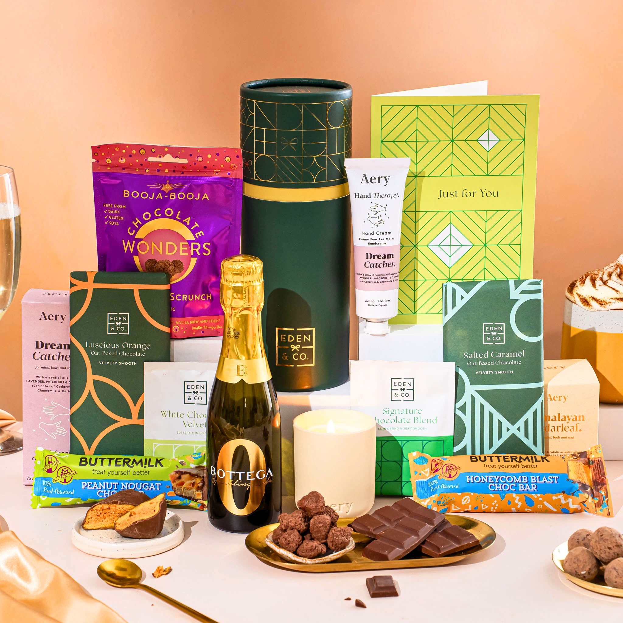 EDEN & CO. plant-based gift hamper with a mini Bottega Zero Black prosecco, green gift tube, “Occasion Themed” card, chocolate bars in Luscious Orange, White Chocolate Velvet, Signature Chocolate Blend and Salted Caramel, Booja-Booja Chocolate Wonders truffles, Buttermilk Peanut Nougat and Honeycomb Blast bars, chocolate truffles and honeycomb pieces on small plates, and a filled champagne glass,  luxury candle and hand cream set against a warm terracotta background.