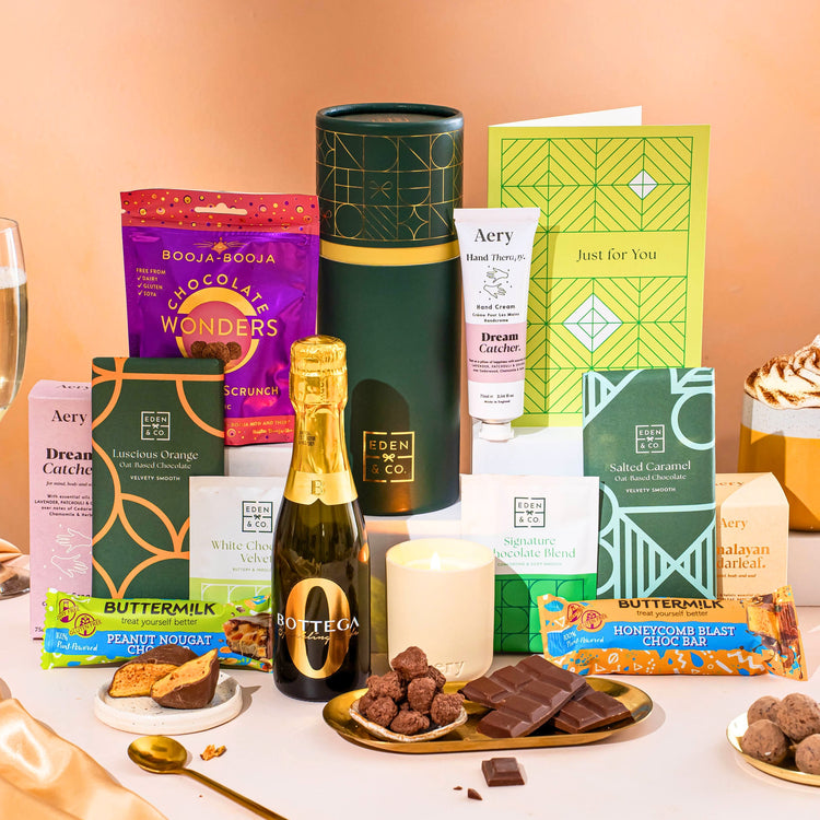 EDEN & CO. plant-based gift hamper with a mini Bottega Zero Black prosecco, green gift tube, “Occasion Themed” card, chocolate bars in Luscious Orange, White Chocolate Velvet, Signature Chocolate Blend and Salted Caramel, Booja-Booja Chocolate Wonders truffles, Buttermilk Peanut Nougat and Honeycomb Blast bars, chocolate truffles and honeycomb pieces on small plates, and a filled champagne glass,  luxury candle and hand cream set against a warm terracotta background.
