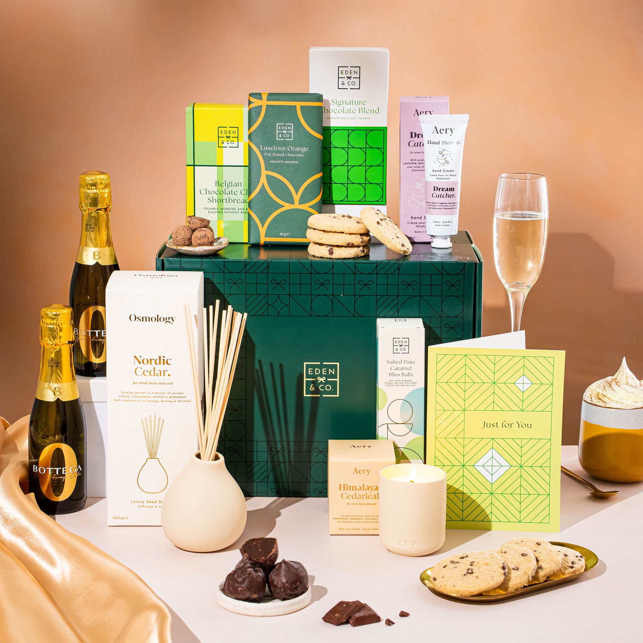 EDEN & CO. Pamper Hamper gift set with a mini Bottega Zero Black prosecco (0% alcohol-free), dark green gift cylinder, an “Occasion Themed” card, chocolate bars and truffles, a scented candle and Aery hand cream, styled with gold silk against a warm terracotta background.
