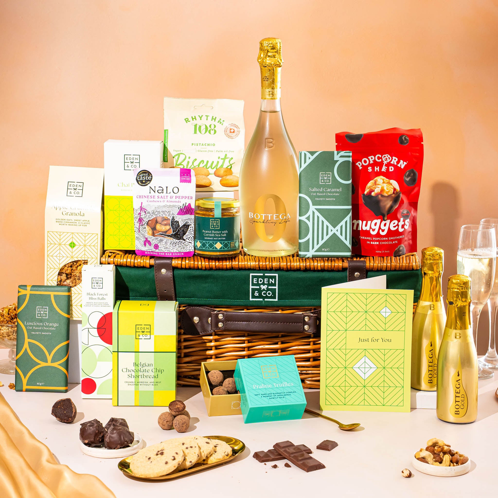EDEN & CO. Luxury Wicker Hamper filled with a full-size Bottega Gold prosecco, an assortment of chocolate bars, Booja-Booja Chocolate Wonders truffles and Buttermilk snack bars, presented in a dark green trimmed wicker basket with an “Occasion Themed” card, styled against a warm terracotta background.