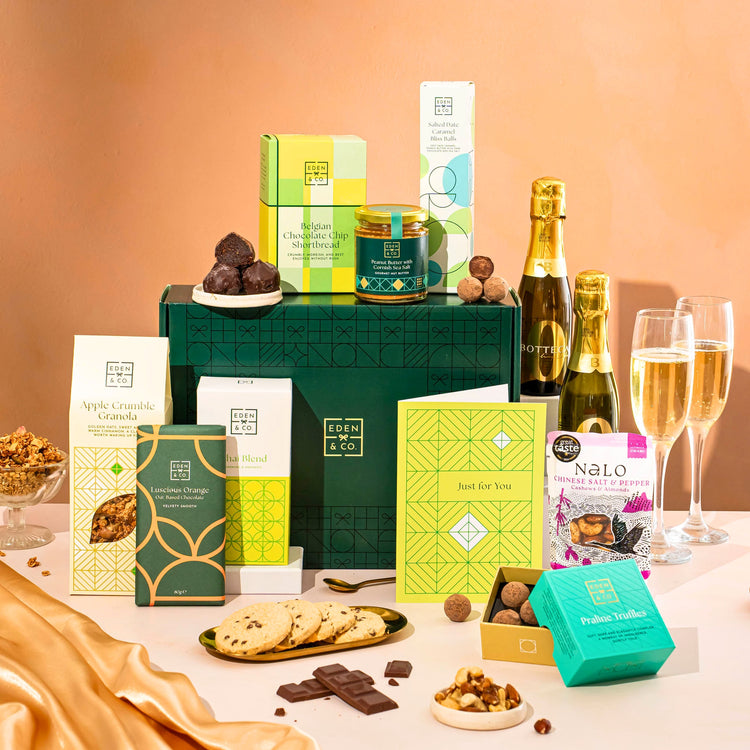 EDEN & CO. Food & Drink Hamper with a full-size Bottega Zero Black prosecco (0% alcohol-free), dark green gift cylinder, an “Occasion Themed” card, an assortment of chocolate bars, Booja-Booja Chocolate Wonders truffles, Buttermilk snack bars, a scented candle and Aery hand cream, styled with gold silk against a warm terracotta background.