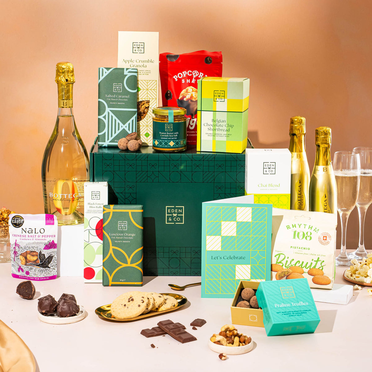 EDEN & CO. Grand Hamper gift box with a full-size Bottega Gold prosecco, dark green gift cylinder, an “Occasion Themed” card, an assortment of chocolate bars, Booja-Booja Chocolate Wonders truffles and Buttermilk snack bars, styled with gold silk against a warm terracotta background.