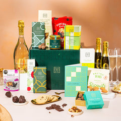 EDEN & CO. Grand Hamper gift box with a full-size Bottega Zero Black prosecco (0% alcohol-free), dark green gift cylinder, an “Occasion Themed” card, an assortment of chocolate bars, Booja-Booja Chocolate Wonders truffles and Buttermilk snack bars, styled with gold silk against a warm terracotta background.