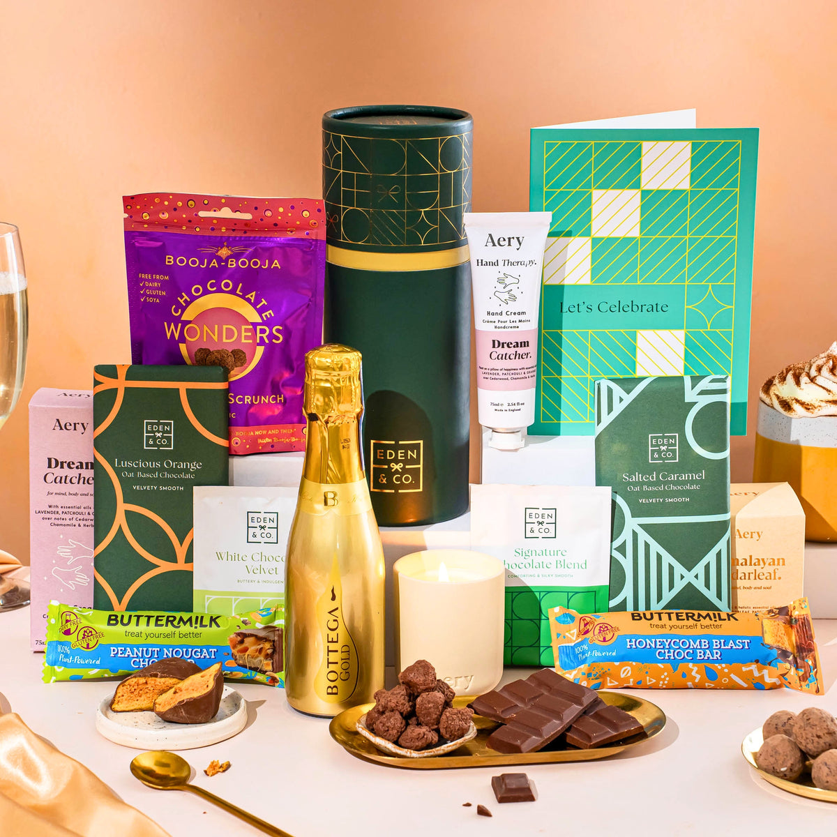 EDEN & CO. plant-based gift hamper with a mini Bottega Gold prosecco, green gift tube, “Occasion Themed” card, chocolate bars in Luscious Orange, White Chocolate Velvet, Signature Chocolate Blend and Salted Caramel, Booja-Booja Chocolate Wonders truffles, Buttermilk Peanut Nougat and Honeycomb Blast bars, chocolate truffles and honeycomb pieces on small plates, and a filled champagne glass, luxury candle and hand cream set against a warm terracotta background.