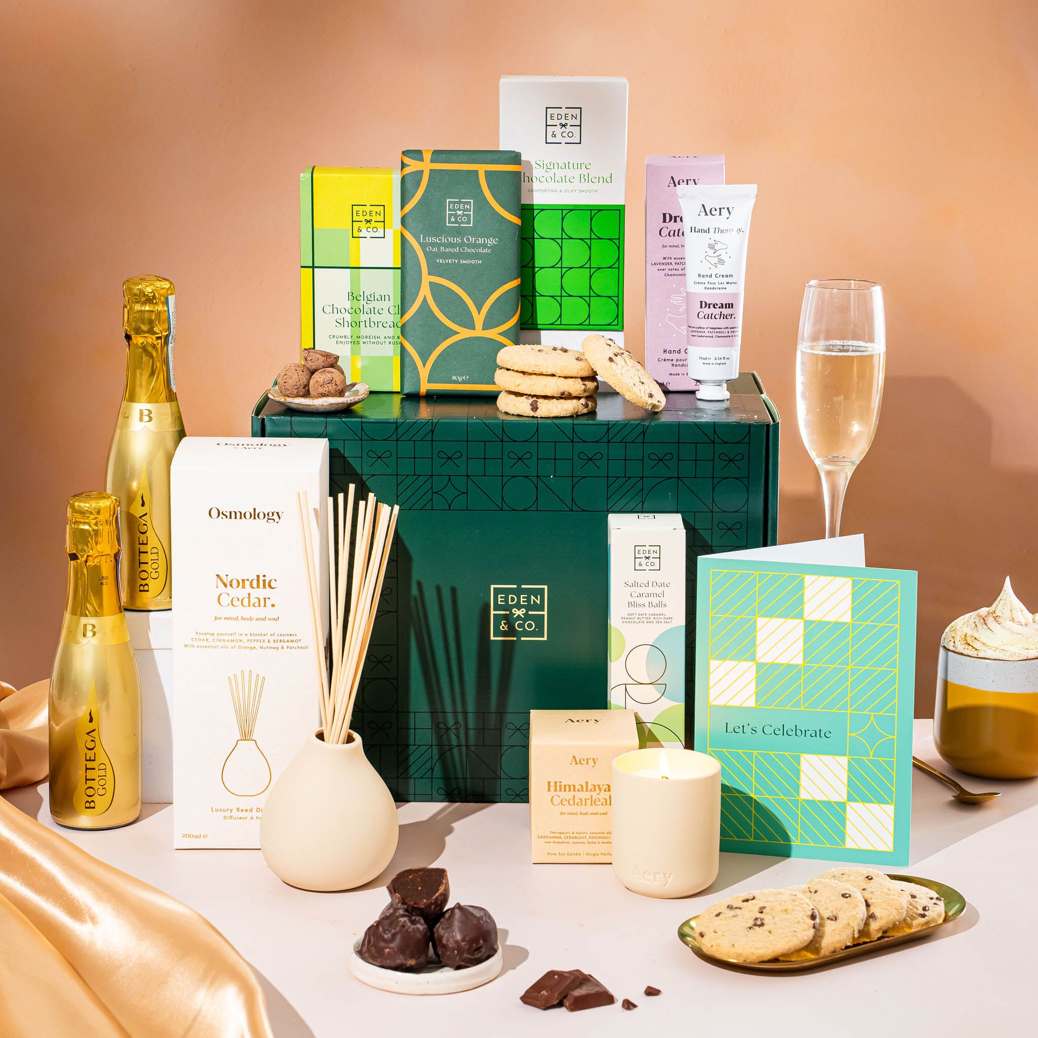 EDEN & CO. Pamper Hamper gift set with a mini Bottega Gold prosecco, dark green gift cylinder, an “Occasion Themed” card, chocolate bars and truffles, a scented candle and Aery hand cream, styled with gold silk against a warm terracotta background.
