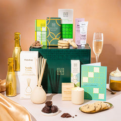 EDEN & CO. Pamper Hamper gift set with a mini Bottega Gold prosecco, dark green gift cylinder, an “Occasion Themed” card, chocolate bars and truffles, a scented candle and Aery hand cream, styled with gold silk against a warm terracotta background.