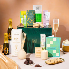 EDEN & CO. Pamper Hamper gift set with a mini Bottega Zero Black prosecco (0% alcohol-free), dark green gift cylinder, an “Occasion Themed” card, chocolate bars and truffles, a scented candle and Aery hand cream, styled with gold silk against a warm terracotta background.
