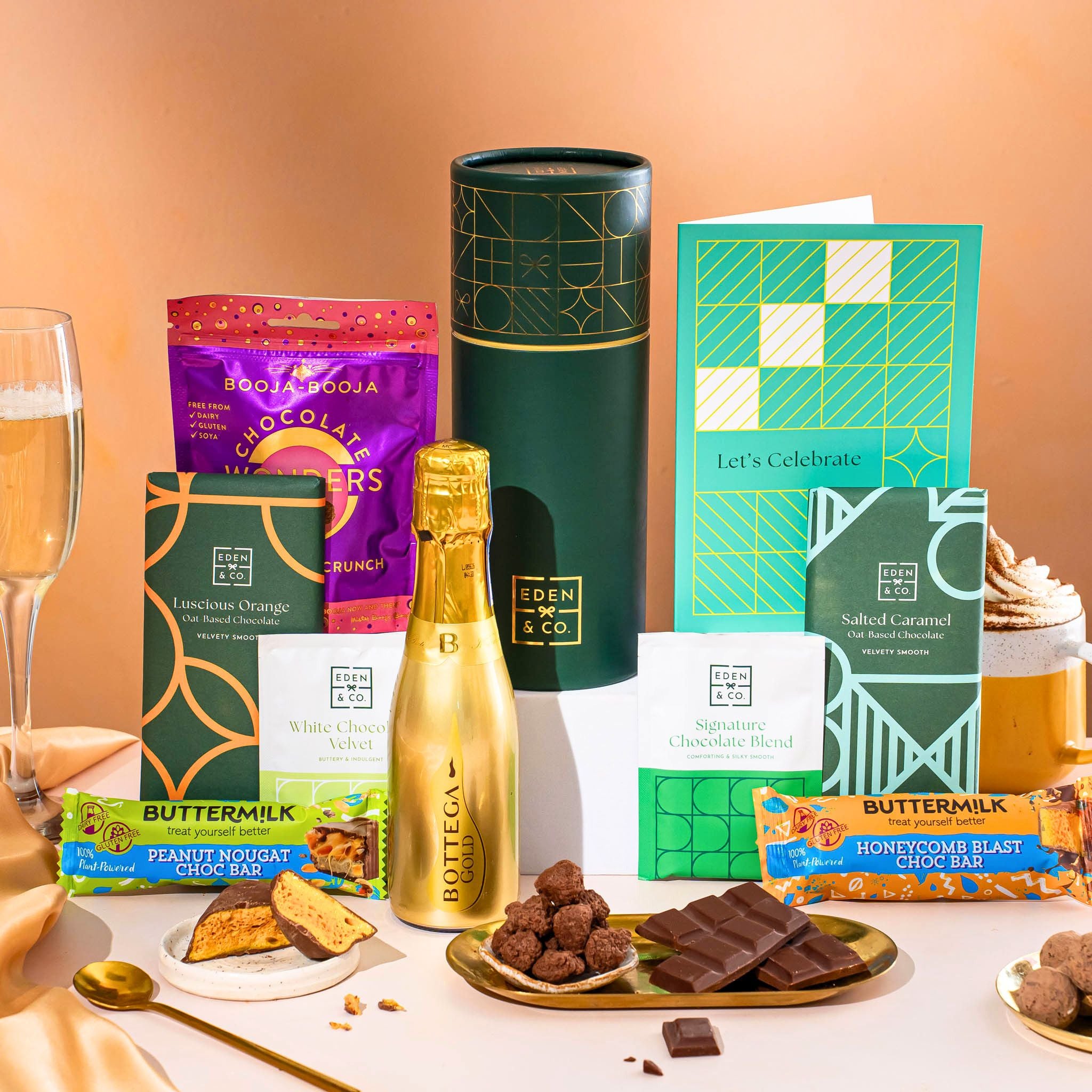 EDEN & CO. plant-based gift hamper with a mini Bottega Gold prosecco, green gift tube, “Occasion Themed” card, chocolate bars in Luscious Orange, White Chocolate Velvet, Signature Chocolate Blend and Salted Caramel, Booja-Booja Chocolate Wonders truffles, Buttermilk Peanut Nougat and Honeycomb Blast bars, chocolate truffles and honeycomb pieces on small plates, and a filled champagne glass, set against a warm terracotta background.