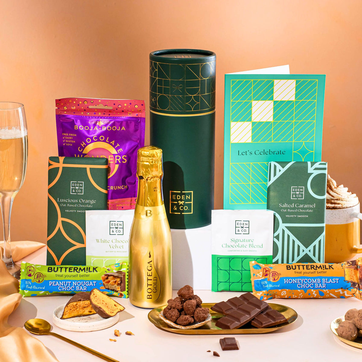 EDEN & CO. plant-based gift hamper with a mini Bottega Gold prosecco, green gift tube, “Occasion Themed” card, chocolate bars in Luscious Orange, White Chocolate Velvet, Signature Chocolate Blend and Salted Caramel, Booja-Booja Chocolate Wonders truffles, Buttermilk Peanut Nougat and Honeycomb Blast bars, chocolate truffles and honeycomb pieces on small plates, and a filled champagne glass, set against a warm terracotta background.