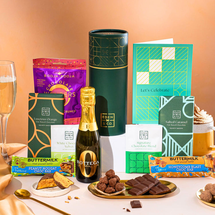 EDEN & CO. plant-based gift hamper with a mini Bottega Zero Black prosecco, green gift tube, “Occasion Themed” card, chocolate bars in Luscious Orange, White Chocolate Velvet, Signature Chocolate Blend and Salted Caramel, Booja-Booja Chocolate Wonders truffles, Buttermilk Peanut Nougat and Honeycomb Blast bars, chocolate truffles and honeycomb pieces on small plates, and a filled champagne glass, set against a warm terracotta background.