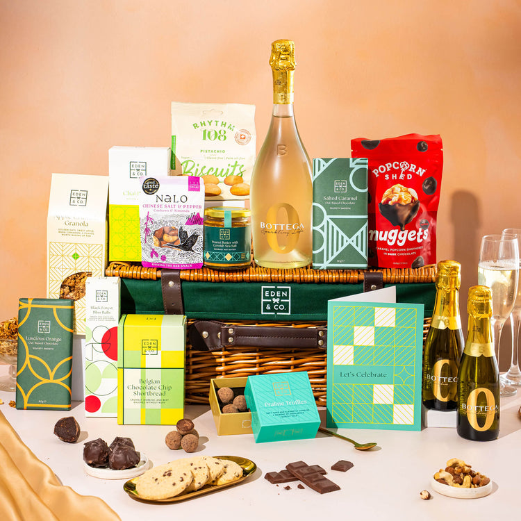 EDEN & CO. Luxury Wicker Hamper filled with a full-size Bottega Zero Black prosecco (0% alcohol-free), an assortment of chocolate bars, Booja-Booja Chocolate Wonders truffles and Buttermilk snack bars, presented in a dark green trimmed wicker basket with an “Occasion Themed” card, styled against a warm terracotta background.