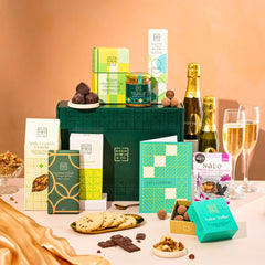 EDEN & CO. Food & Drink Hamper with a full-size Bottega Zero Black prosecco (0% alcohol-free), dark green gift cylinder, an “Occasion Themed” card, an assortment of chocolate bars, Booja-Booja Chocolate Wonders truffles, Buttermilk snack bars, a scented candle and Aery hand cream, styled with gold silk against a warm terracotta background.