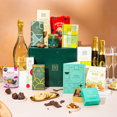 EDEN & CO. Grand Hamper gift box with a full-size Bottega Zero Black prosecco (0% alcohol-free), dark green gift cylinder, an “Occasion Themed” card, an assortment of chocolate bars, Booja-Booja Chocolate Wonders truffles and Buttermilk snack bars, styled with gold silk against a warm terracotta background.