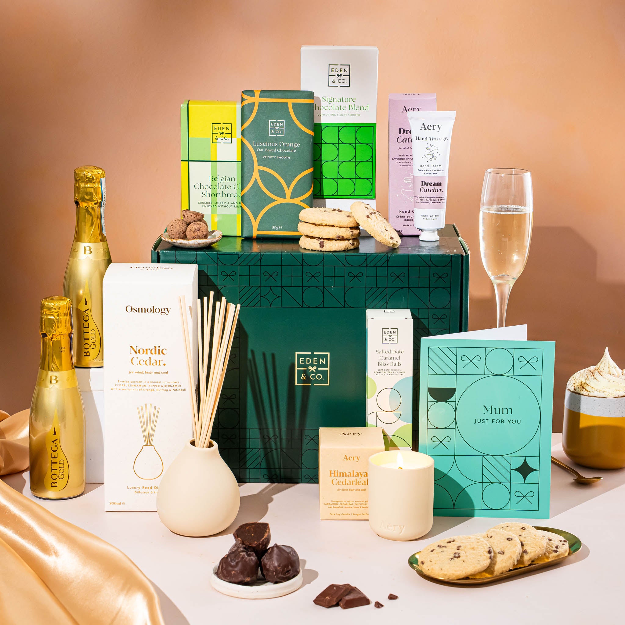 EDEN & CO. Pamper Hamper gift set with a mini Bottega Gold prosecco, dark green gift cylinder, an “Occasion Themed” card, chocolate bars and truffles, a scented candle and Aery hand cream, styled with gold silk against a warm terracotta background.