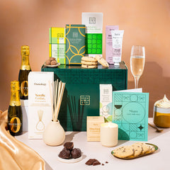 EDEN & CO. Pamper Hamper gift set with a mini Bottega Zero Black prosecco (0% alcohol-free), dark green gift cylinder, an “Occasion Themed” card, chocolate bars and truffles, a scented candle and Aery hand cream, styled with gold silk against a warm terracotta background.