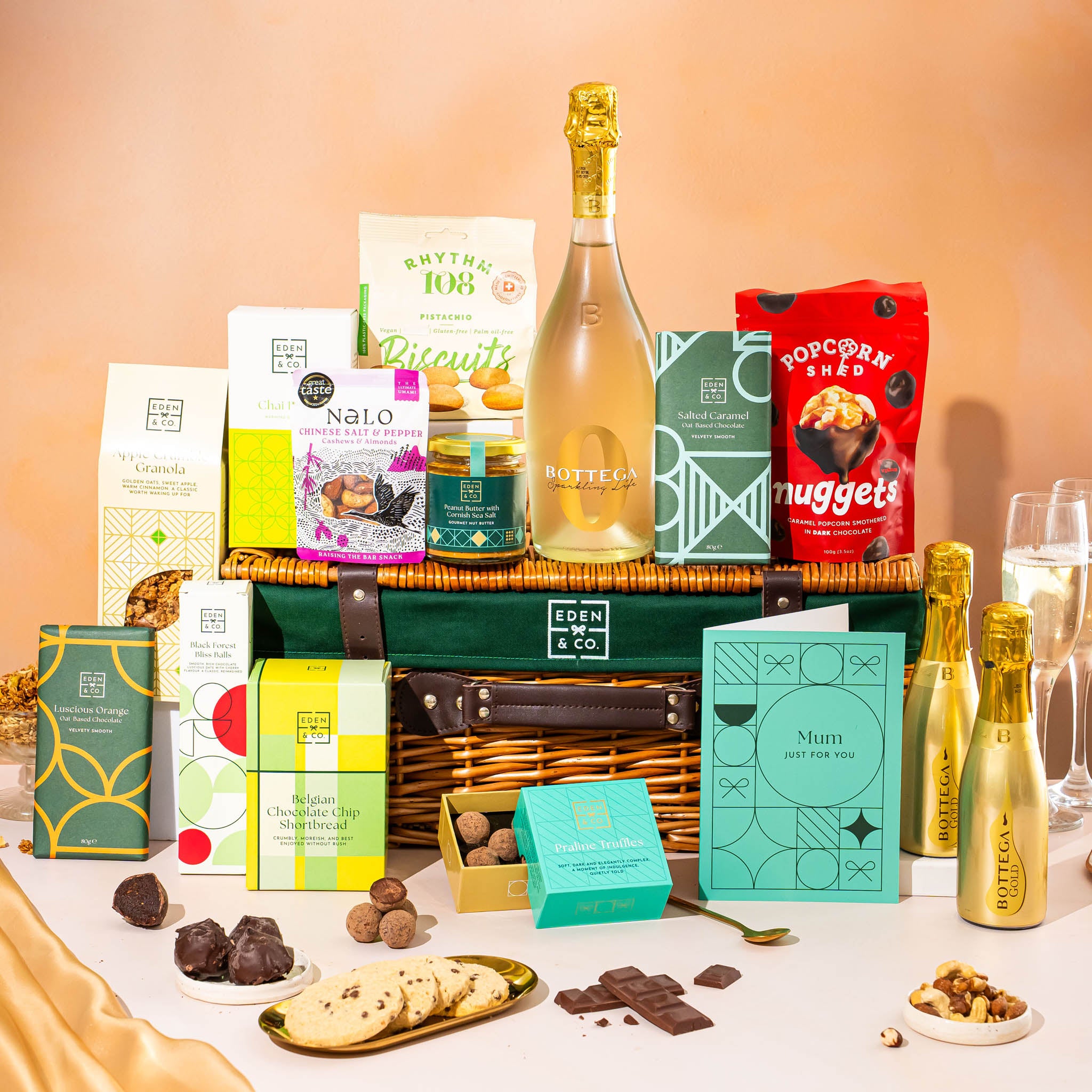 EDEN & CO. Luxury Wicker Hamper filled with a full-size Bottega Gold prosecco, an assortment of chocolate bars, Booja-Booja Chocolate Wonders truffles and Buttermilk snack bars, presented in a dark green trimmed wicker basket with an “Occasion Themed” card, styled against a warm terracotta background.