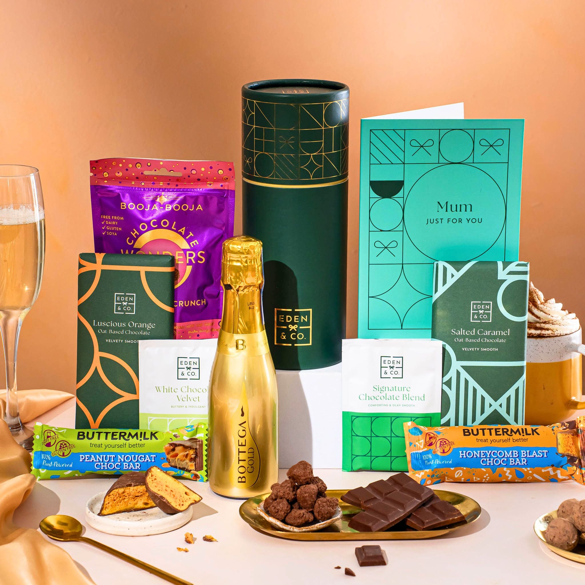 EDEN & CO. plant-based gift hamper with a mini Bottega Gold prosecco, green gift tube, “Occasion Themed” card, chocolate bars in Luscious Orange, White Chocolate Velvet, Signature Chocolate Blend and Salted Caramel, Booja-Booja Chocolate Wonders truffles, Buttermilk Peanut Nougat and Honeycomb Blast bars, chocolate truffles and honeycomb pieces on small plates, and a filled champagne glass, set against a warm terracotta background.