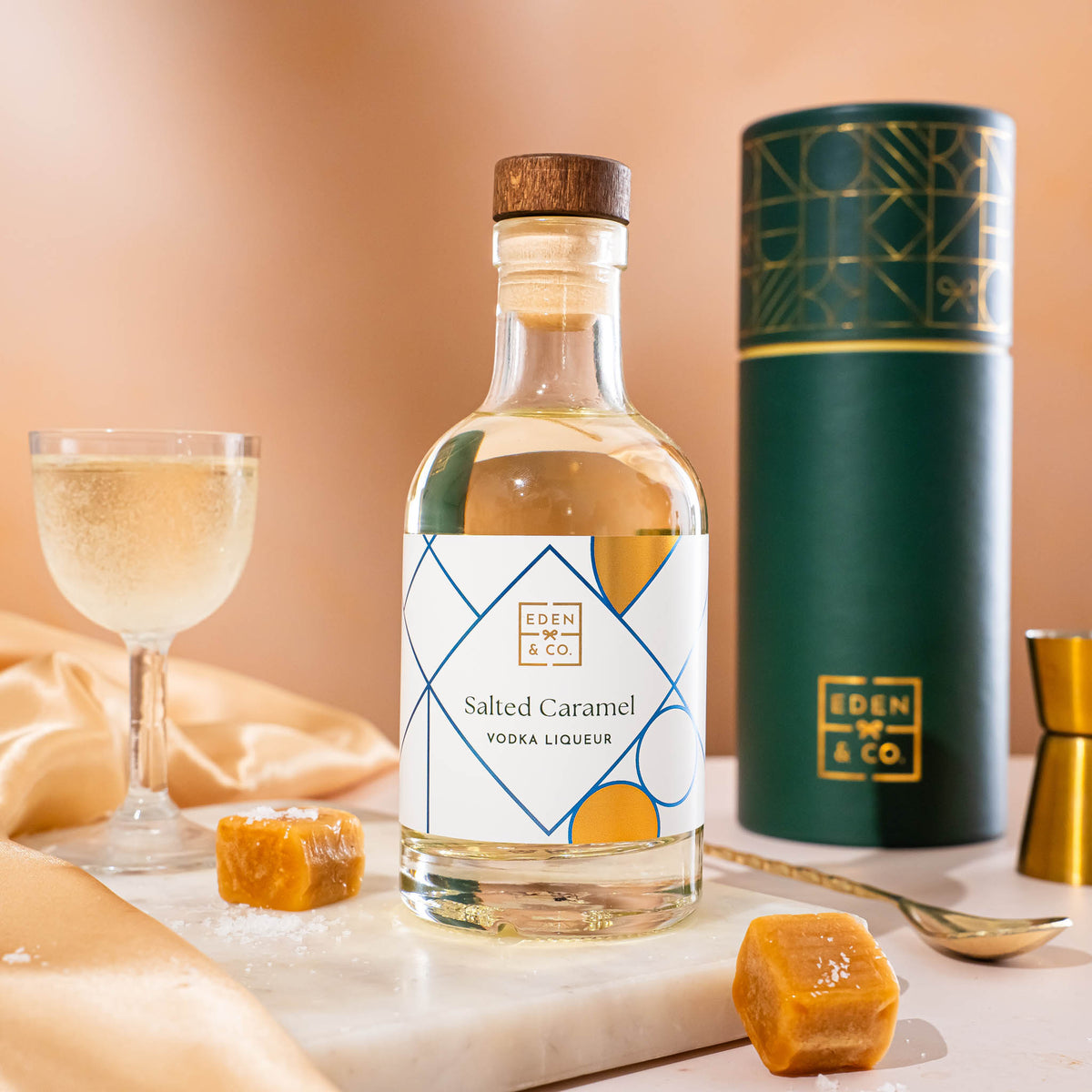 EDEN & CO. liqueur gift bottle with an “Occasion Themed” card, presented in a branded gift tube, styled with gold silk against a warm terracotta background.