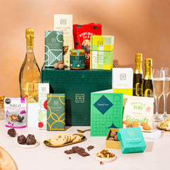 EDEN & CO. Grand Hamper gift box with a full-size Bottega Zero Black prosecco (0% alcohol-free), dark green gift cylinder, an “Occasion Themed” card, an assortment of chocolate bars, Booja-Booja Chocolate Wonders truffles and Buttermilk snack bars, styled with gold silk against a warm terracotta background.