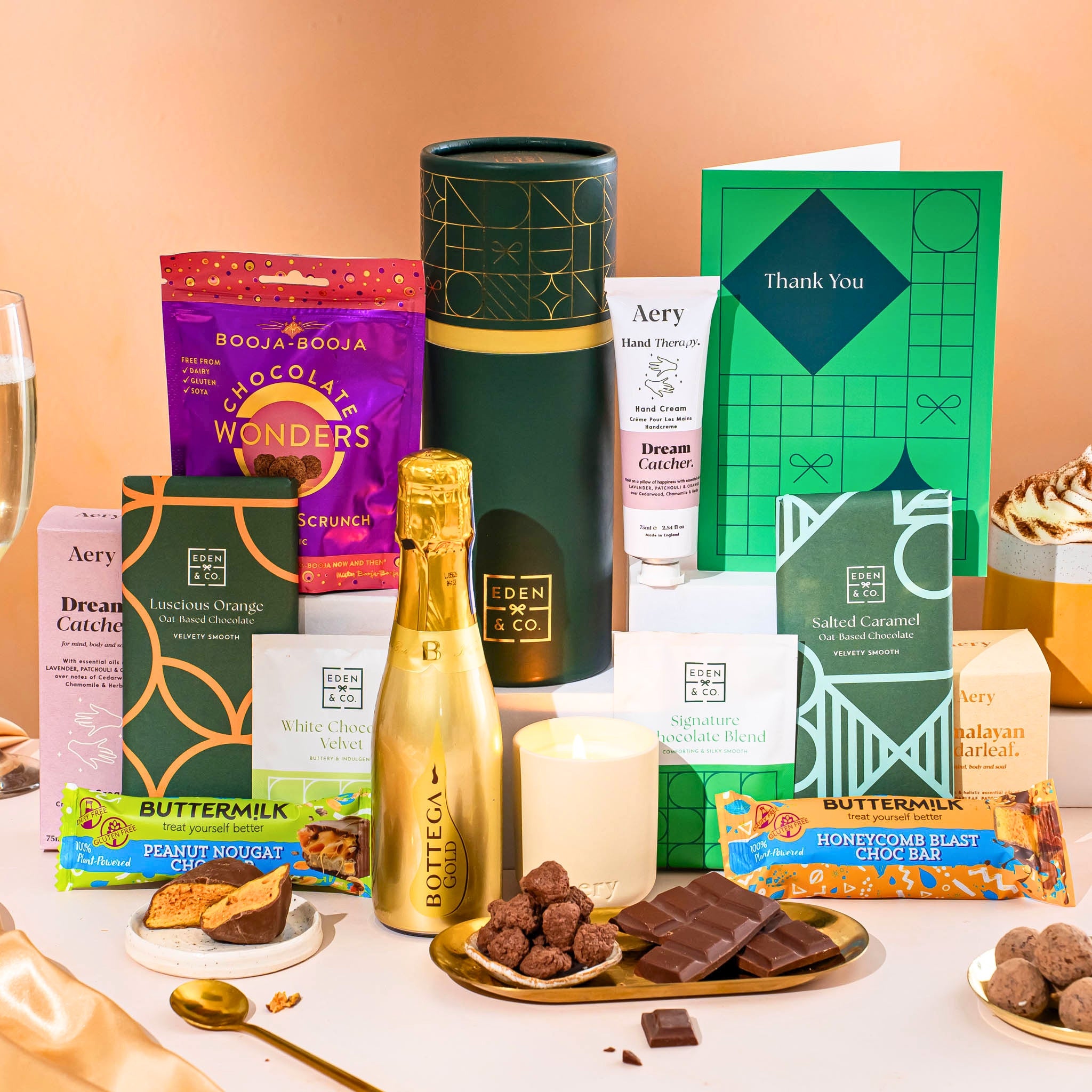 EDEN & CO. plant-based gift hamper with a mini Bottega Gold prosecco, green gift tube, “Occasion Themed” card, chocolate bars in Luscious Orange, White Chocolate Velvet, Signature Chocolate Blend and Salted Caramel, Booja-Booja Chocolate Wonders truffles, Buttermilk Peanut Nougat and Honeycomb Blast bars, chocolate truffles and honeycomb pieces on small plates, and a filled champagne glass, luxury candle and hand cream set against a warm terracotta background.