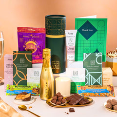 EDEN & CO. plant-based gift hamper with a mini Bottega Gold prosecco, green gift tube, “Occasion Themed” card, chocolate bars in Luscious Orange, White Chocolate Velvet, Signature Chocolate Blend and Salted Caramel, Booja-Booja Chocolate Wonders truffles, Buttermilk Peanut Nougat and Honeycomb Blast bars, chocolate truffles and honeycomb pieces on small plates, and a filled champagne glass, luxury candle and hand cream set against a warm terracotta background.