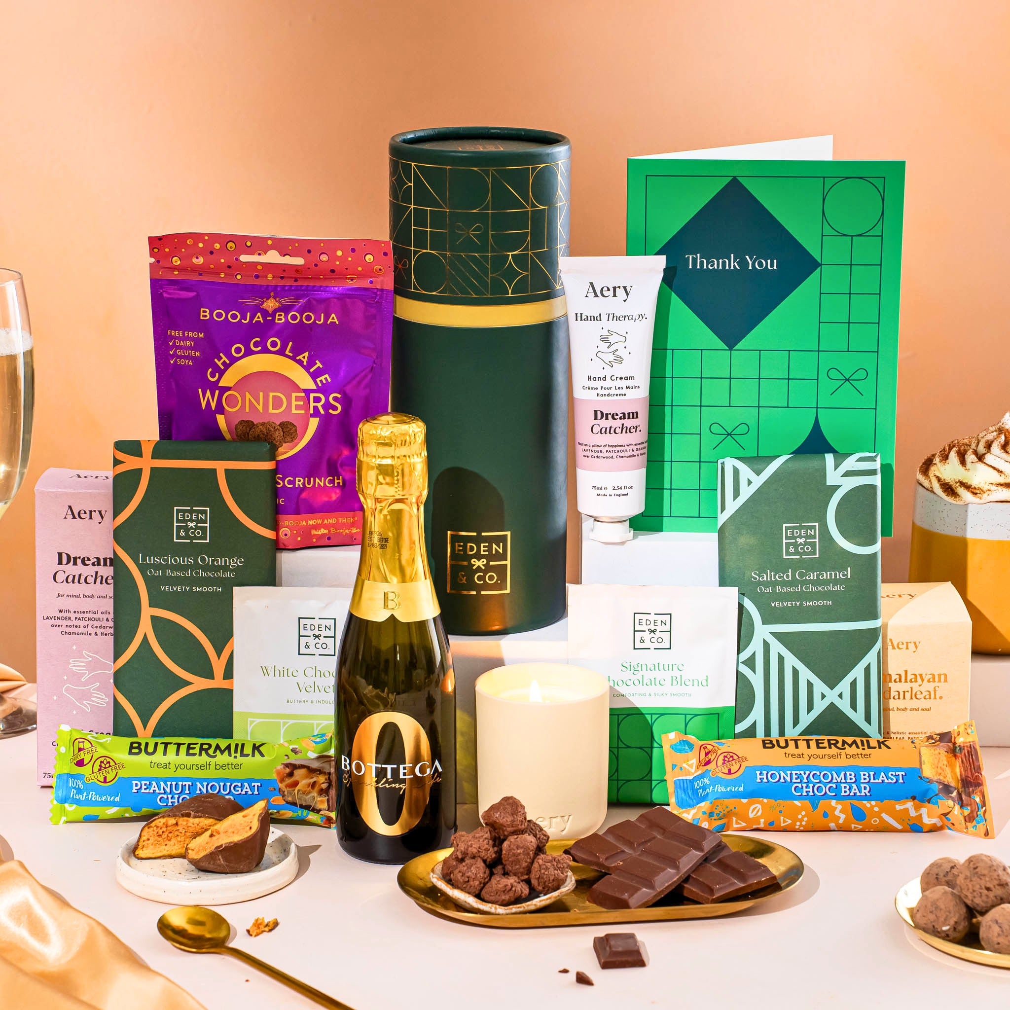 EDEN & CO. plant-based gift hamper with a mini Bottega Zero Black prosecco, green gift tube, “Occasion Themed” card, chocolate bars in Luscious Orange, White Chocolate Velvet, Signature Chocolate Blend and Salted Caramel, Booja-Booja Chocolate Wonders truffles, Buttermilk Peanut Nougat and Honeycomb Blast bars, chocolate truffles and honeycomb pieces on small plates, and a filled champagne glass,  luxury candle and hand cream set against a warm terracotta background.