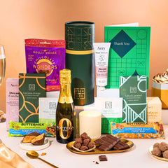 EDEN & CO. plant-based gift hamper with a mini Bottega Zero Black prosecco, green gift tube, “Occasion Themed” card, chocolate bars in Luscious Orange, White Chocolate Velvet, Signature Chocolate Blend and Salted Caramel, Booja-Booja Chocolate Wonders truffles, Buttermilk Peanut Nougat and Honeycomb Blast bars, chocolate truffles and honeycomb pieces on small plates, and a filled champagne glass,  luxury candle and hand cream set against a warm terracotta background.