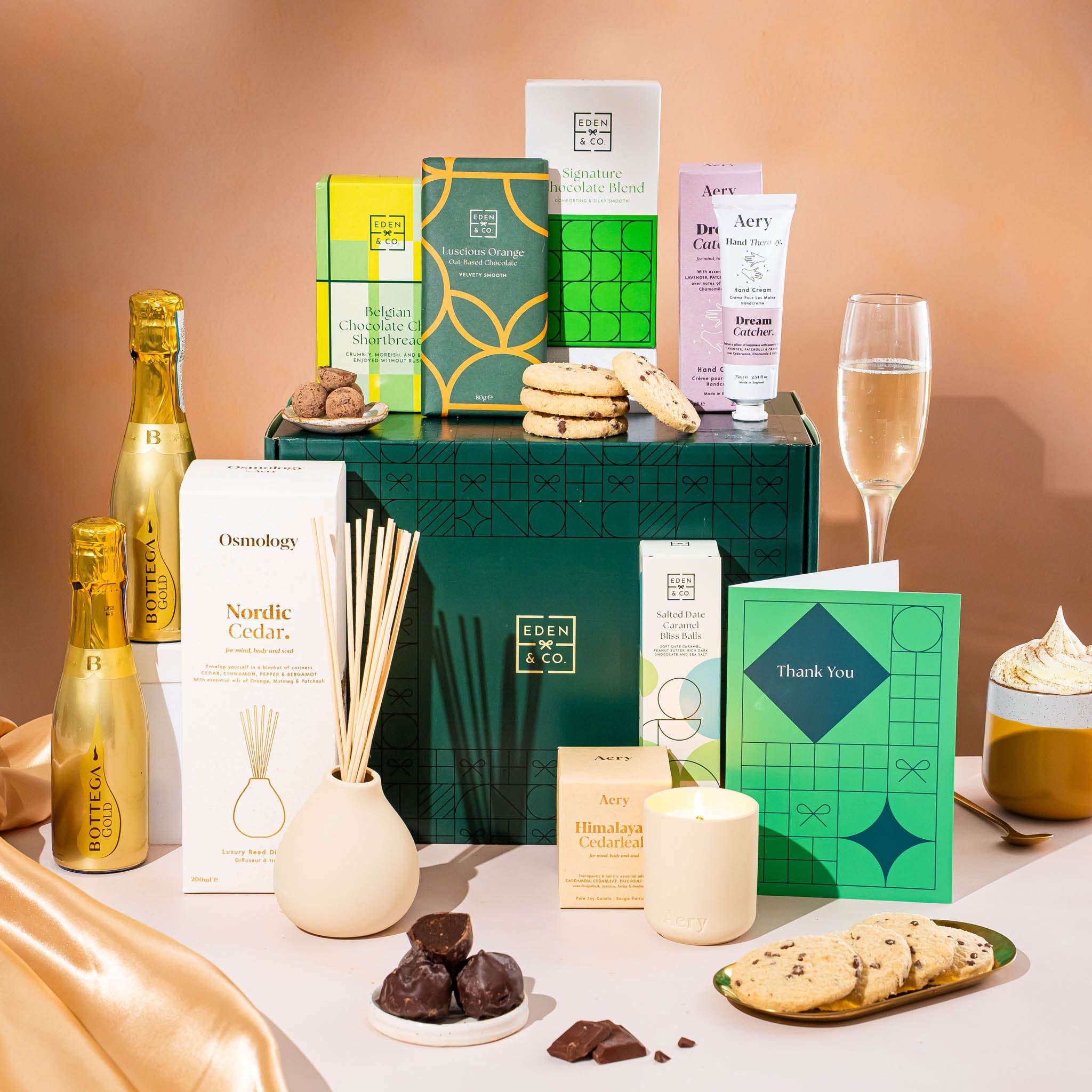 EDEN & CO. Pamper Hamper gift set with a mini Bottega Gold prosecco, dark green gift cylinder, an “Occasion Themed” card, chocolate bars and truffles, a scented candle and Aery hand cream, styled with gold silk against a warm terracotta background.