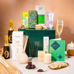 EDEN & CO. Pamper Hamper gift set with a mini Bottega Zero Black prosecco (0% alcohol-free), dark green gift cylinder, an “Occasion Themed” card, chocolate bars and truffles, a scented candle and Aery hand cream, styled with gold silk against a warm terracotta background.