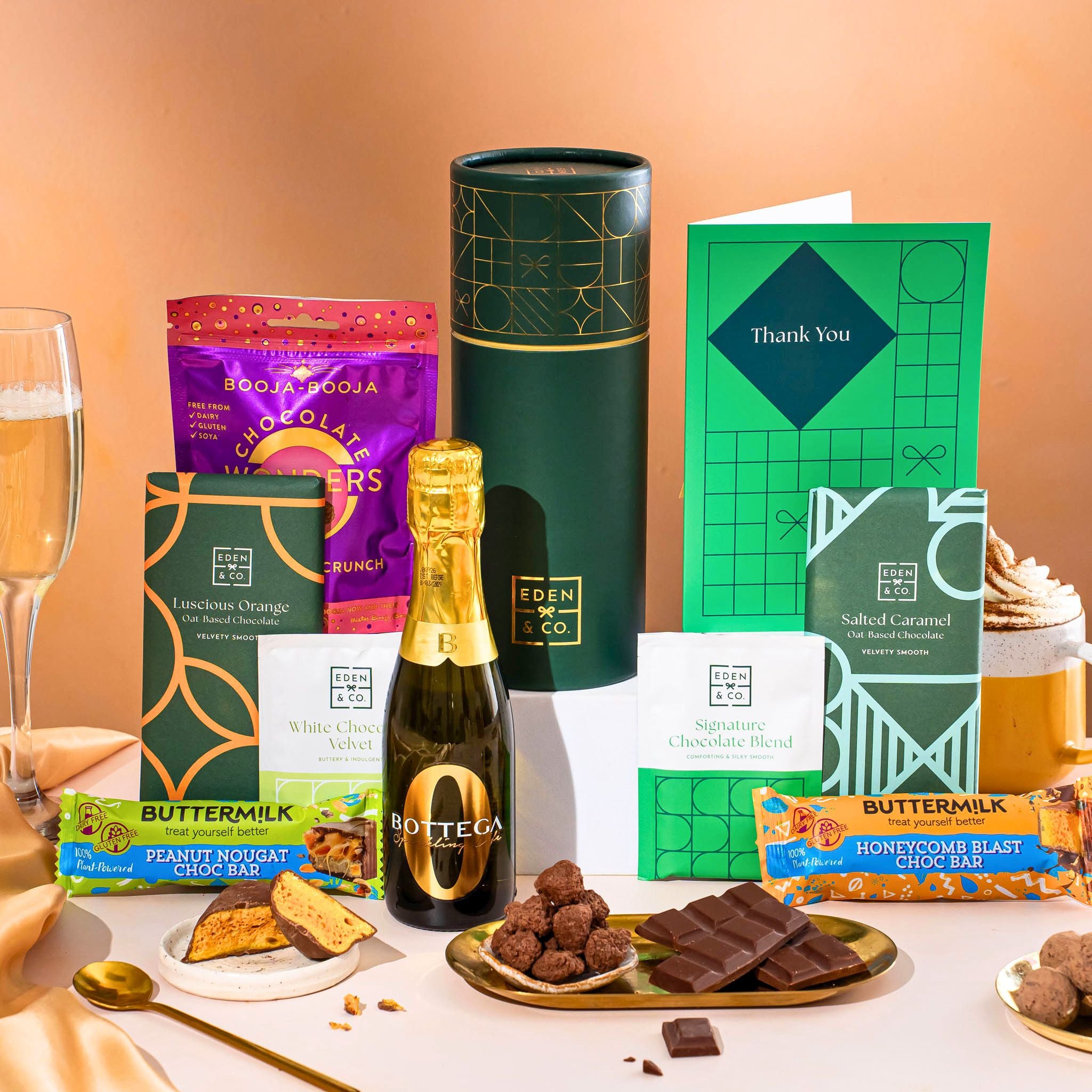 EDEN & CO. plant-based gift hamper with a mini Bottega Zero Black prosecco, green gift tube, “Occasion Themed” card, chocolate bars in Luscious Orange, White Chocolate Velvet, Signature Chocolate Blend and Salted Caramel, Booja-Booja Chocolate Wonders truffles, Buttermilk Peanut Nougat and Honeycomb Blast bars, chocolate truffles and honeycomb pieces on small plates, and a filled champagne glass, set against a warm terracotta background.
