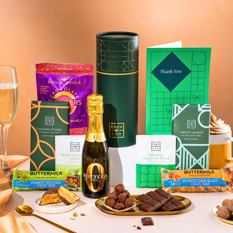 EDEN & CO. plant-based gift hamper with a mini Bottega Zero Black prosecco, green gift tube, “Occasion Themed” card, chocolate bars in Luscious Orange, White Chocolate Velvet, Signature Chocolate Blend and Salted Caramel, Booja-Booja Chocolate Wonders truffles, Buttermilk Peanut Nougat and Honeycomb Blast bars, chocolate truffles and honeycomb pieces on small plates, and a filled champagne glass, set against a warm terracotta background.