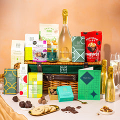 EDEN & CO. Luxury Wicker Hamper filled with a full-size Bottega Gold prosecco, an assortment of chocolate bars, Booja-Booja Chocolate Wonders truffles and Buttermilk snack bars, presented in a dark green trimmed wicker basket with an “Occasion Themed” card, styled against a warm terracotta background.