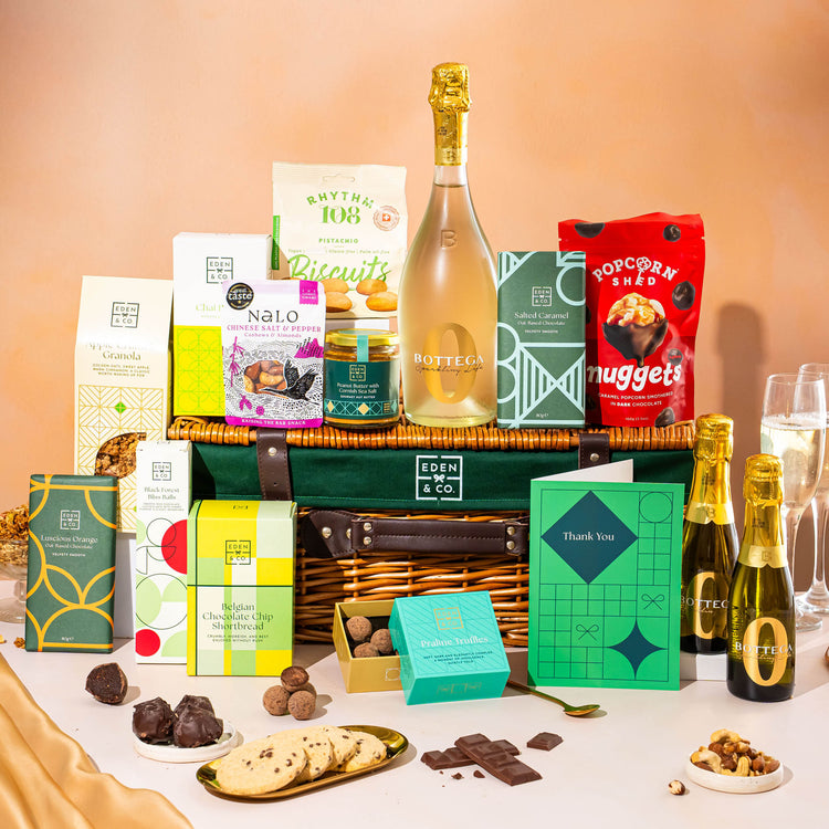 EDEN & CO. Luxury Wicker Hamper filled with a full-size Bottega Zero Black prosecco (0% alcohol-free), an assortment of chocolate bars, Booja-Booja Chocolate Wonders truffles and Buttermilk snack bars, presented in a dark green trimmed wicker basket with an “Occasion Themed” card, styled against a warm terracotta background.