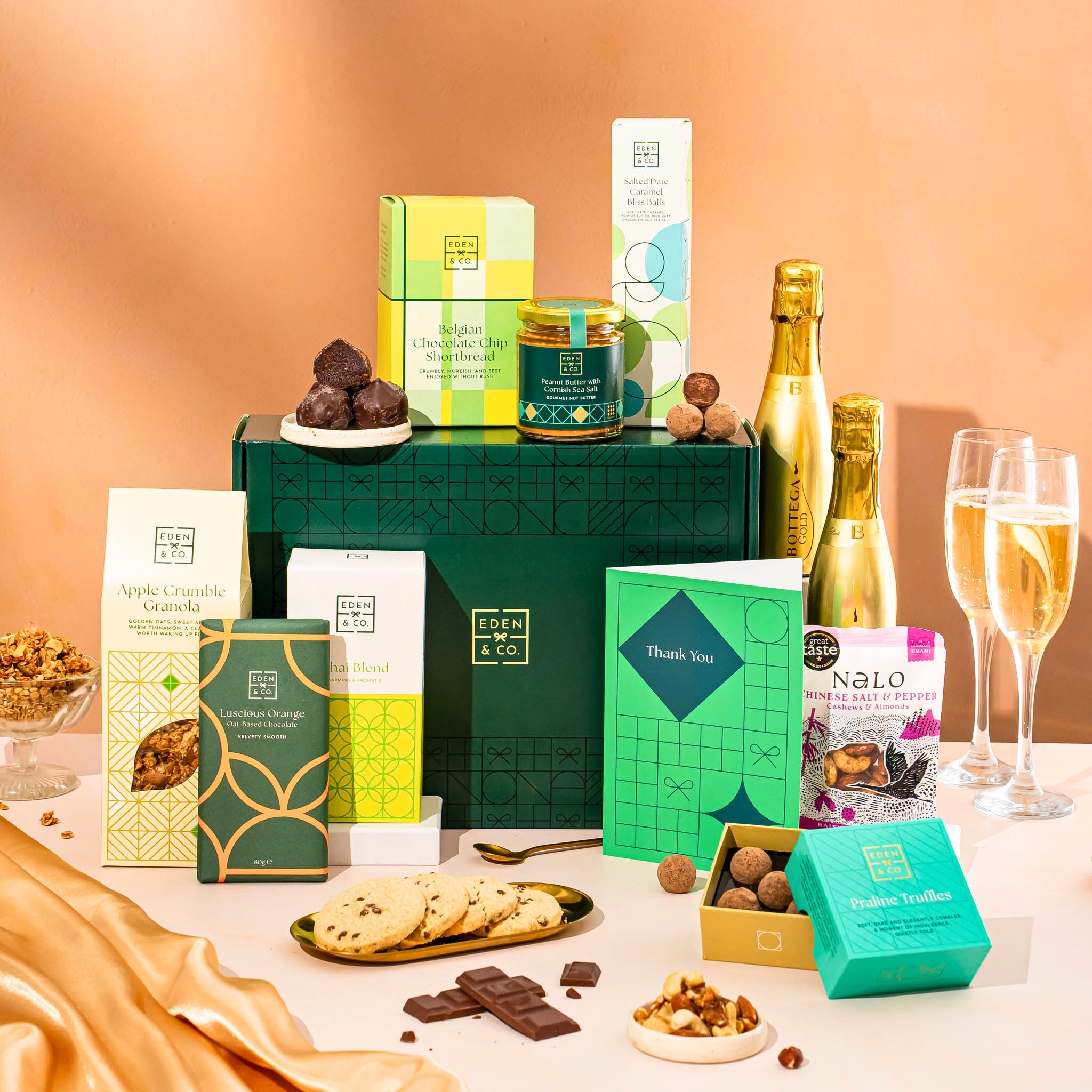 EDEN & CO. Food & Drink Hamper with a full-size Bottega Gold prosecco, dark green gift cylinder, an “Occasion Themed” card, an assortment of chocolate bars, Booja-Booja Chocolate Wonders truffles, Buttermilk snack bars, a scented candle and Aery hand cream, styled with gold silk against a warm terracotta background.