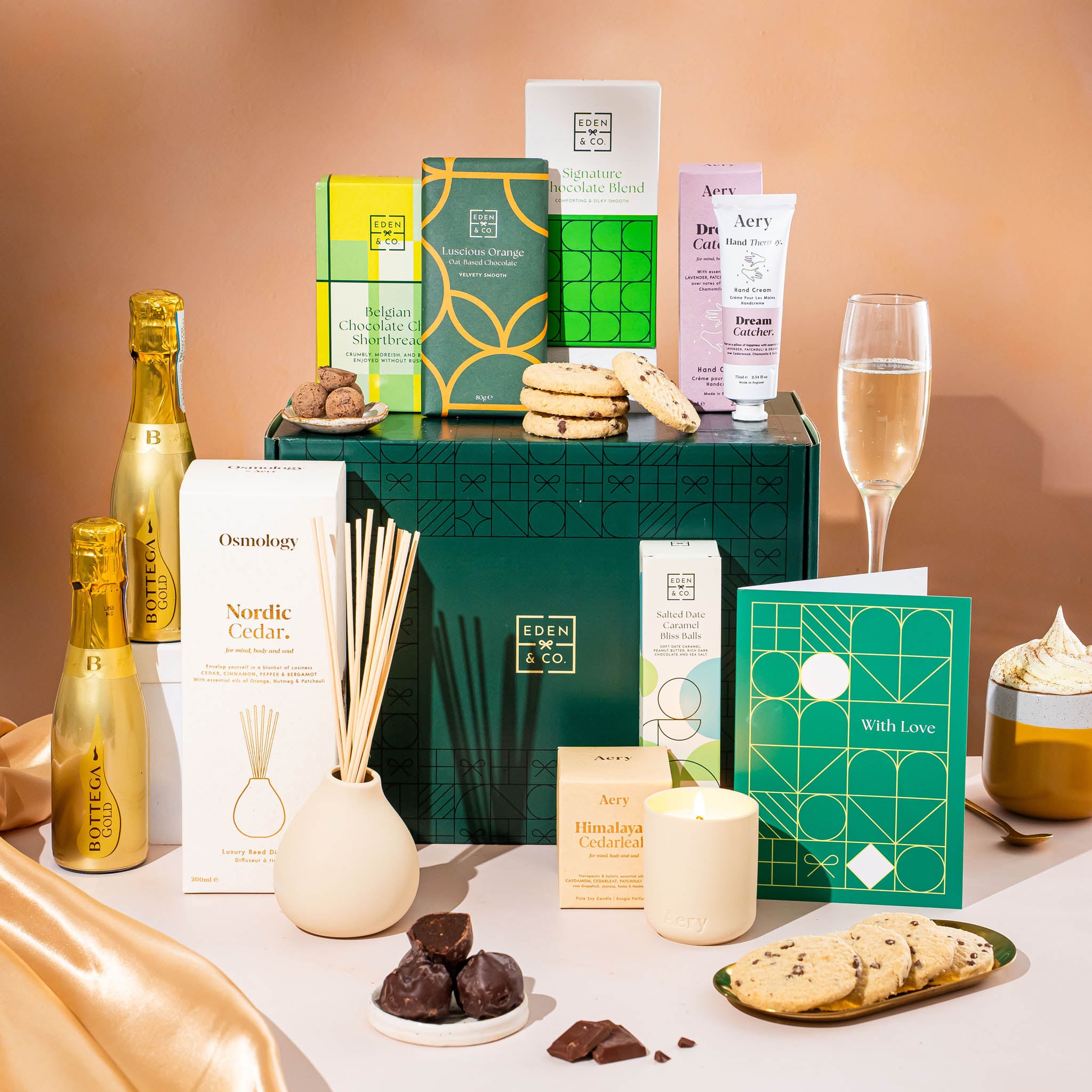 EDEN & CO. Pamper Hamper gift set with a mini Bottega Gold prosecco, dark green gift cylinder, an “Occasion Themed” card, chocolate bars and truffles, a scented candle and Aery hand cream, styled with gold silk against a warm terracotta background.