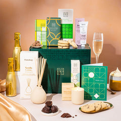 EDEN & CO. Pamper Hamper gift set with a mini Bottega Gold prosecco, dark green gift cylinder, an “Occasion Themed” card, chocolate bars and truffles, a scented candle and Aery hand cream, styled with gold silk against a warm terracotta background.