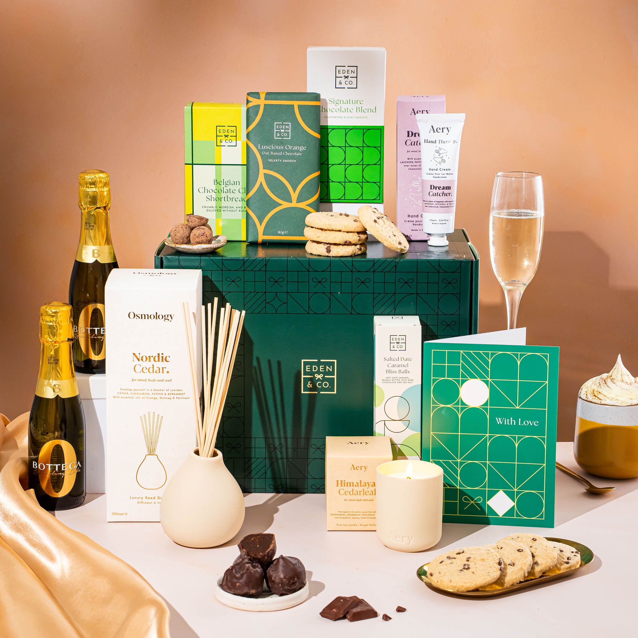EDEN & CO. Pamper Hamper gift set with a mini Bottega Zero Black prosecco (0% alcohol-free), dark green gift cylinder, an “Occasion Themed” card, chocolate bars and truffles, a scented candle and Aery hand cream, styled with gold silk against a warm terracotta background.