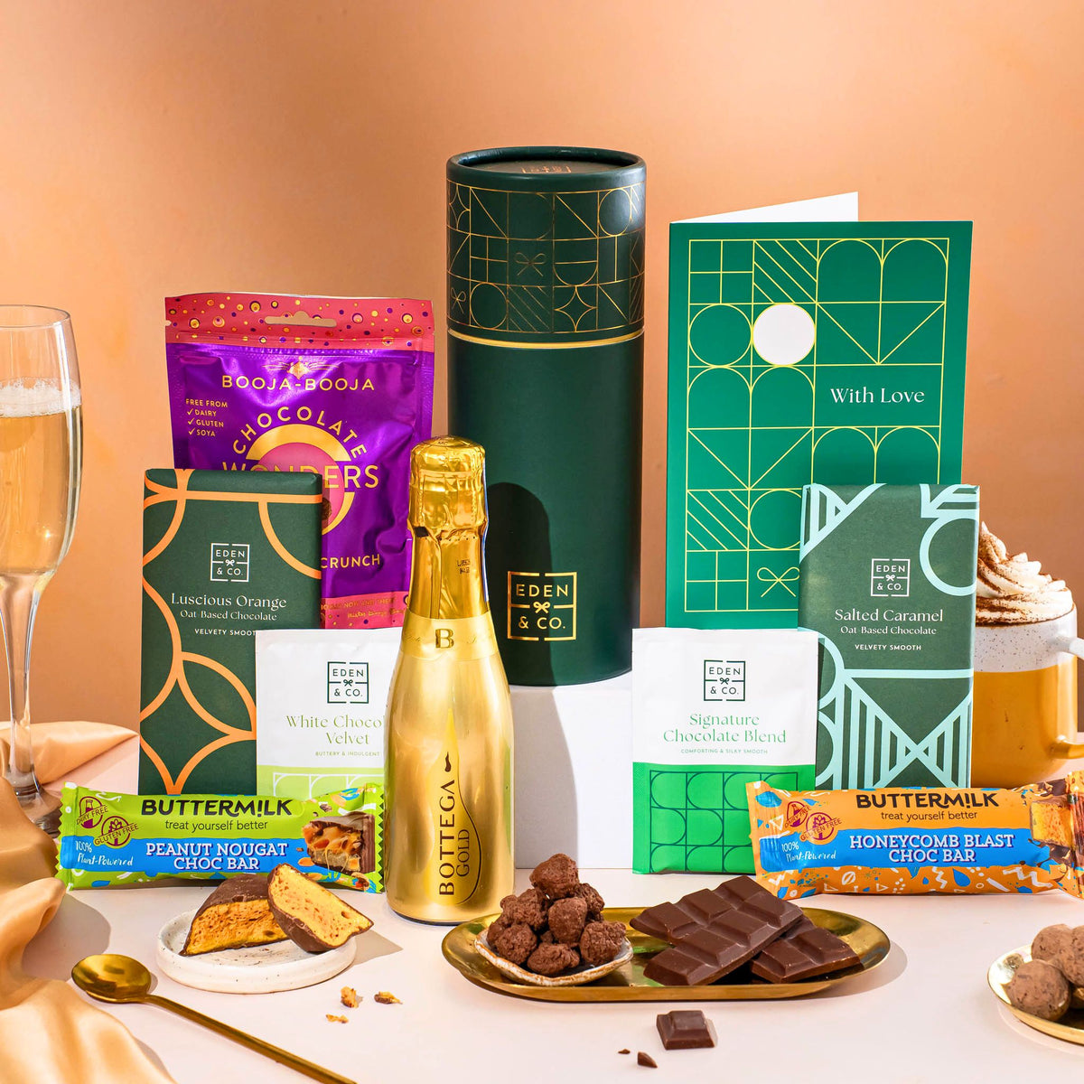 EDEN & CO. plant-based gift hamper with a mini Bottega Gold prosecco, green gift tube, “Occasion Themed” card, chocolate bars in Luscious Orange, White Chocolate Velvet, Signature Chocolate Blend and Salted Caramel, Booja-Booja Chocolate Wonders truffles, Buttermilk Peanut Nougat and Honeycomb Blast bars, chocolate truffles and honeycomb pieces on small plates, and a filled champagne glass, set against a warm terracotta background.