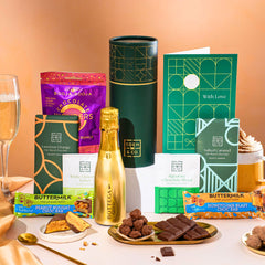 EDEN & CO. plant-based gift hamper with a mini Bottega Gold prosecco, green gift tube, “Occasion Themed” card, chocolate bars in Luscious Orange, White Chocolate Velvet, Signature Chocolate Blend and Salted Caramel, Booja-Booja Chocolate Wonders truffles, Buttermilk Peanut Nougat and Honeycomb Blast bars, chocolate truffles and honeycomb pieces on small plates, and a filled champagne glass, set against a warm terracotta background.