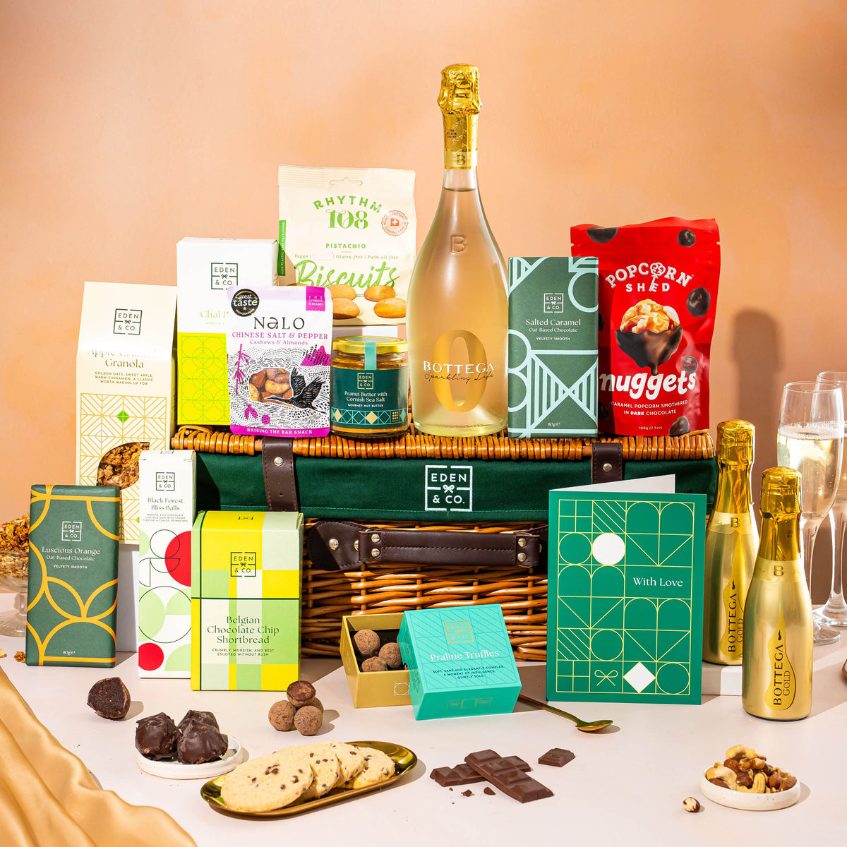 EDEN & CO. Luxury Wicker Hamper filled with a full-size Bottega Gold prosecco, an assortment of chocolate bars, Booja-Booja Chocolate Wonders truffles and Buttermilk snack bars, presented in a dark green trimmed wicker basket with an “Occasion Themed” card, styled against a warm terracotta background.