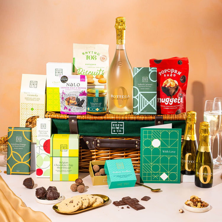 EDEN & CO. Luxury Wicker Hamper filled with a full-size Bottega Zero Black prosecco (0% alcohol-free), an assortment of chocolate bars, Booja-Booja Chocolate Wonders truffles and Buttermilk snack bars, presented in a dark green trimmed wicker basket with an “Occasion Themed” card, styled against a warm terracotta background.