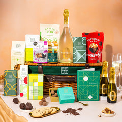 EDEN & CO. Luxury Wicker Hamper filled with a full-size Bottega Zero Black prosecco (0% alcohol-free), an assortment of chocolate bars, Booja-Booja Chocolate Wonders truffles and Buttermilk snack bars, presented in a dark green trimmed wicker basket with an “Occasion Themed” card, styled against a warm terracotta background.