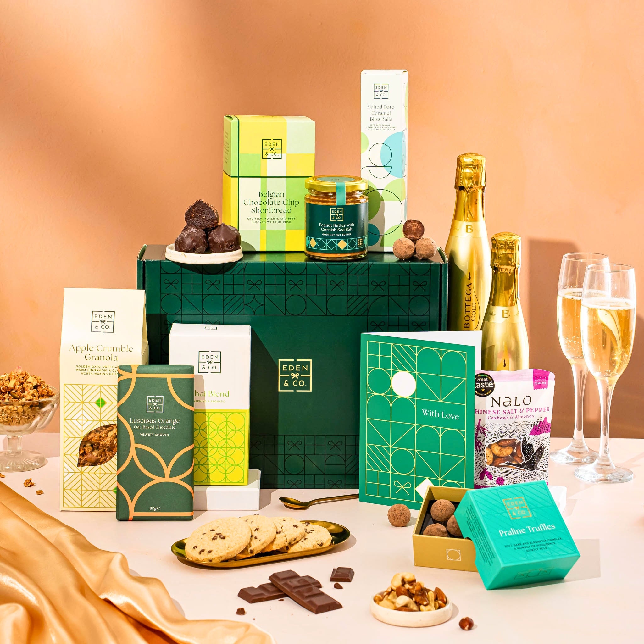 EDEN & CO. Food & Drink Hamper with a full-size Bottega Gold prosecco, dark green gift cylinder, an “Occasion Themed” card, an assortment of chocolate bars, Booja-Booja Chocolate Wonders truffles, Buttermilk snack bars, a scented candle and Aery hand cream, styled with gold silk against a warm terracotta background.