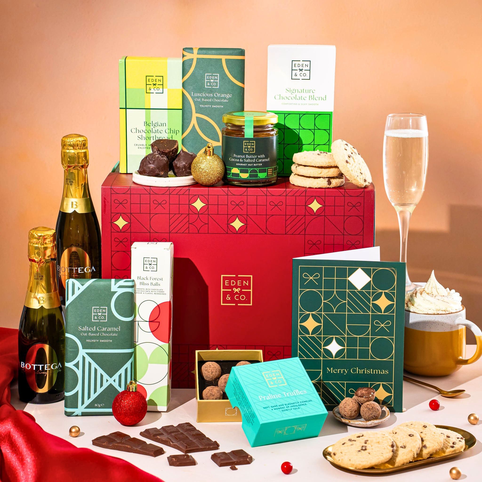 EDEN & CO. Chocolate Hamper letterbox gift with a mini Bottega Zero Black prosecco (0% alcohol-free), green gift tube, an “Occasion Themed” card, an assortment of chocolate bars, Booja-Booja Chocolate Wonders truffles and chocolate truffles on a small plate, styled against a warm terracotta background.